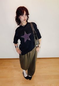 Svart mesh-tröja med en lila stjärngrafik, kombinerad med lösa olivgröna byxor. Accessoarer inkluderar silverarmband och en liten väska.