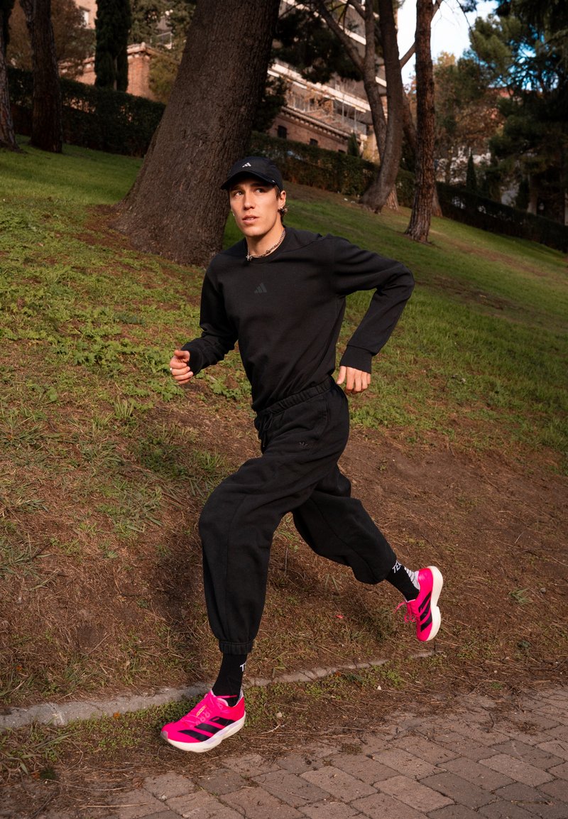 Completo sportivo nero composto da una felpa girocollo e pantaloni da jogging. Scarpe da corsa rosa acceso, valorizzate da calzini neri. Sullo sfondo, erba verde e alberi.