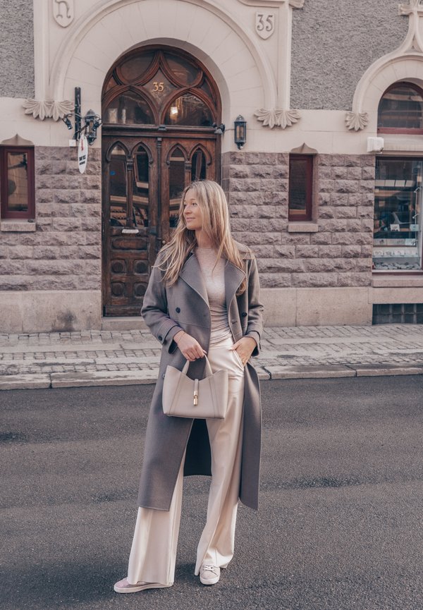 Ženska v dolgem sivem plašču in krem hlačah drži bež torbico, stoji na ulici pred kamnito zgradbo z obokanim lesenim vratom.