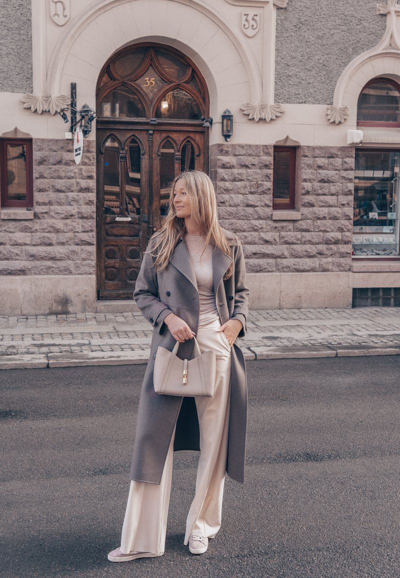 Femme portant un long manteau gris et un pantalon crème, tenant un sac à main beige, debout dans une rue devant un bâtiment en pierre avec une porte en bois arquée.