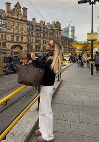 Bruine geruite shopper, zwarte jas, witte wijd uitlopende broek en bruine enkellaarsjes. Stedelijke omgeving met gele leuningen en historische gebouwen.