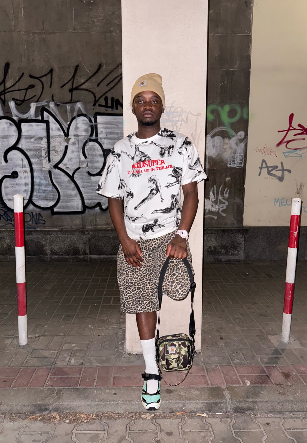 Streetwear outfit featuring a white graphic t-shirt with black and red accents, leopard print shorts, and green-accented trainers. Camo bag.