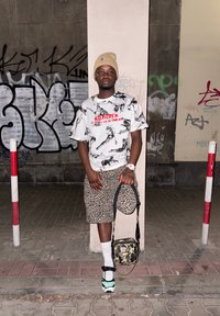 Streetwear outfit featuring a white graphic t-shirt with black and red accents, leopard print shorts, and green-accented trainers. Camo bag.