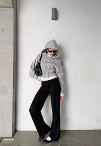 Sweat à capuche gris raccourci avec une fermeture éclair à l'avant, associé à un pantalon noir évasé. La tenue comprend un sac noir et des lunettes de soleil foncées. Textures lisses.