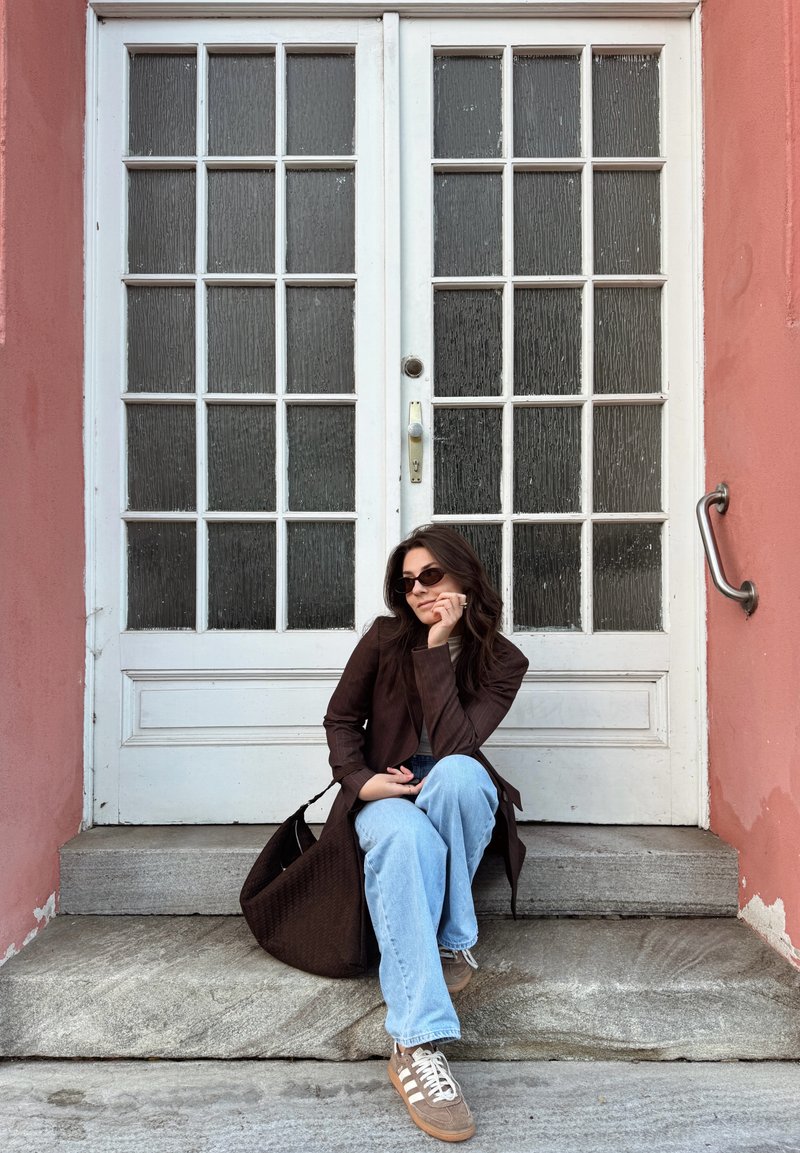 Bruine gestructureerde jas, lichtblauwe wijde jeans, bruine sneakers met witte strepen. Zittend op stenen stappen met een witte dubbele deur op de achtergrond.