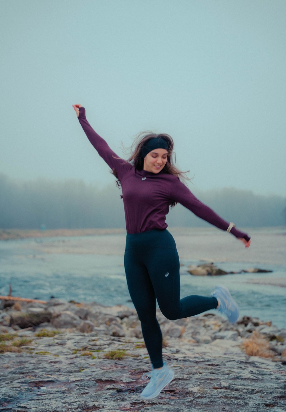 Femme en tenue de sport sautant joyeusement sur une rive rocheuse avec de l'eau et une forêt brumeuse en arrière-plan.