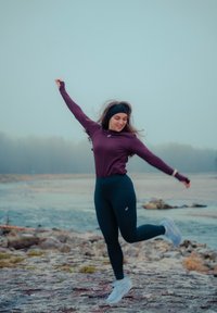 Femme en tenue de sport sautant joyeusement sur une rive rocheuse avec de l'eau et une forêt brumeuse en arrière-plan.