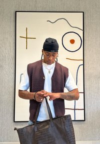 Mann med briller, hodeplagg og tatovert arm som har på seg strikket vest og hvit skjorte, holder en stor svart teksturert totebag innendørs.