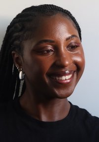 Retrato de una persona con cabello trenzado, que lleva una blusa negra y pendientes de plata. La piel tiene una textura suave, con una iluminación tenue que resalta los rasgos faciales.