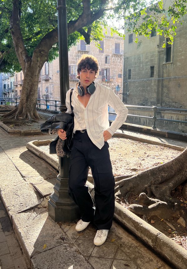 Camicia a righe bianche con bottoni, pantaloni cargo neri, sneakers bianche e cuffie. In piedi vicino a un albero in un'area di ciottoli.