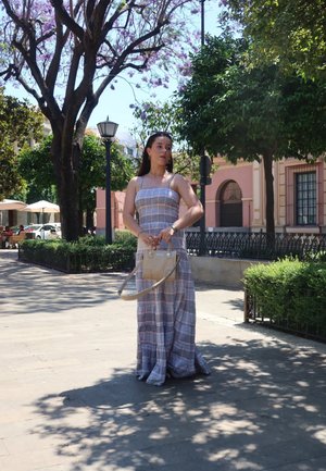 Vestido maxi largo a cuadros en tonos claros con tirantes finos. Combinado con un pequeño bolso beige con correa para el hombro. Ambiente al aire libre con árboles.