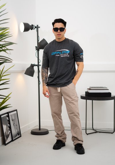 Camiseta gris de gran tamaño con texto azul "OFFLINE", pantalones cargo beige, zapatillas negras y gafas de sol; ambiente interior con planta y lámpara.
