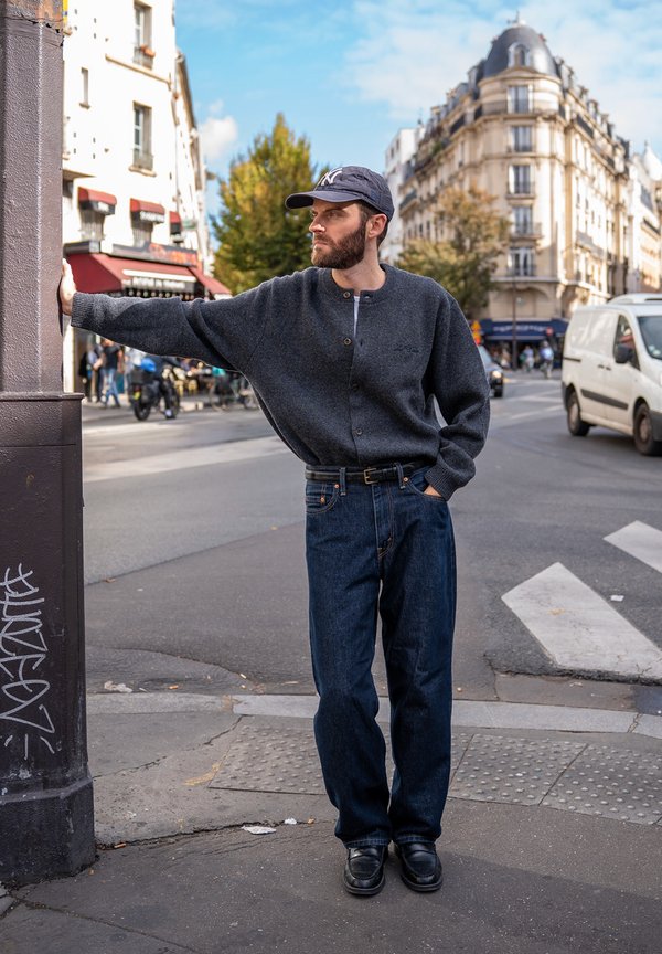 Un hombre con barba, gorra, chaqueta de punto gris y vaqueros se apoya en un poste de la calle en una concurrida esquina de la ciudad.
