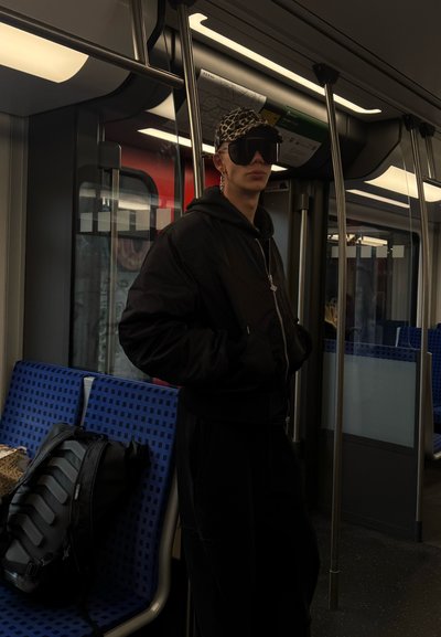 Chaqueta bomber negra, pantalones a juego, gafas de sol de gran tamaño y una gorra de estampado de leopardo. Una mochila negra descansa sobre un asiento de patrón azul en el metro.