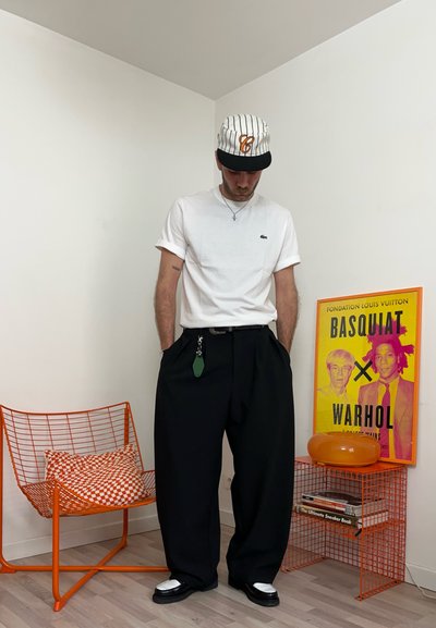 Hombre con camiseta blanca, gorra a rayas y pantalones anchos negros, de pie con las manos en los bolsillos junto a una silla de alambre naranja y un colorido póster de Basquiat Warhol.