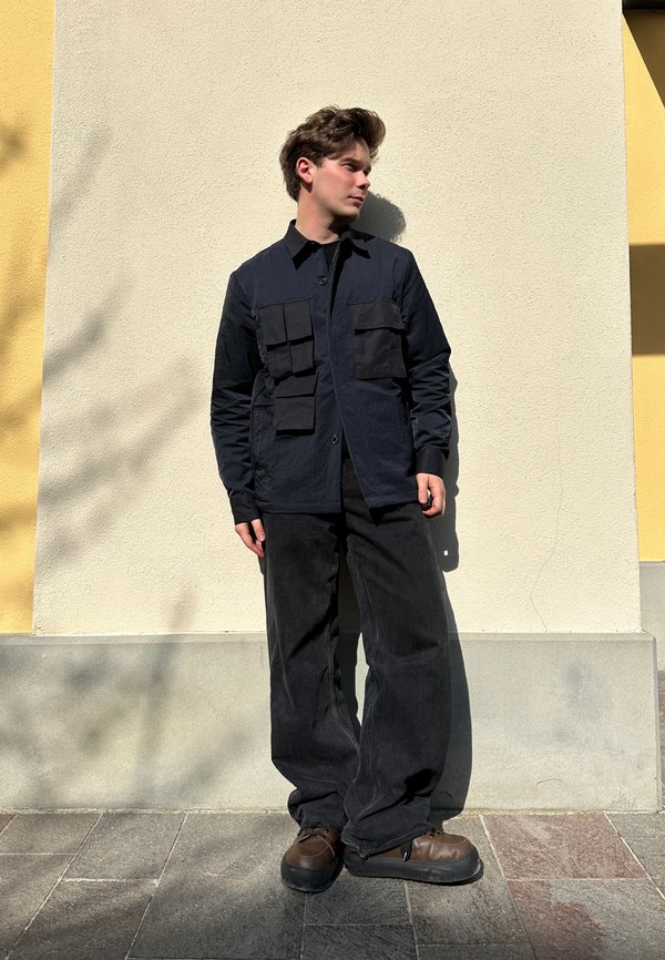 Mørk marineblå knappjakke med fire store lommer, kombinert med løse svarte bukser og brune klumpete sko mot en lys bakgrunn.