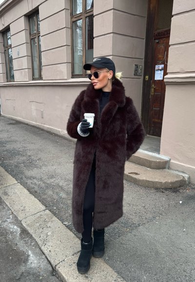 Capa de piel burdeos larga con un collar ancho, llevada sobre un conjunto negro. Guantes negros y botas de tobillo negras gruesas. Sosteniendo una taza.
