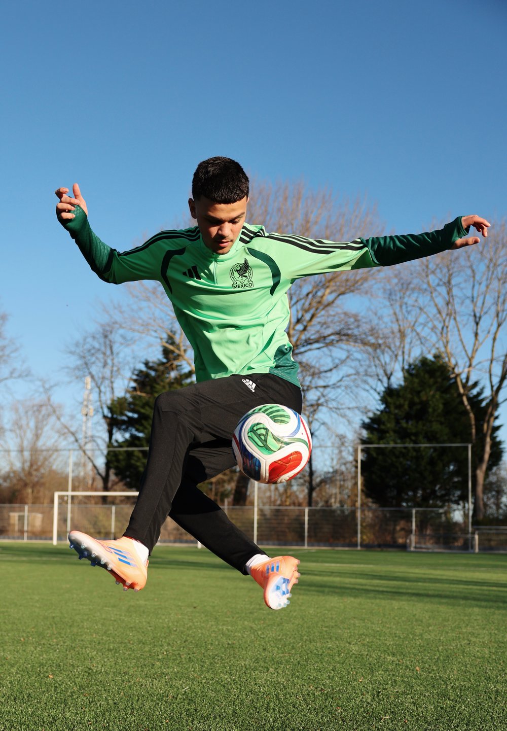 Jeune joueur de football masculin portant un maillot du Mexique, sautant pour frapper un ballon de football sur un terrain herbeux avec des arbres sans feuilles et un ciel dégagé.