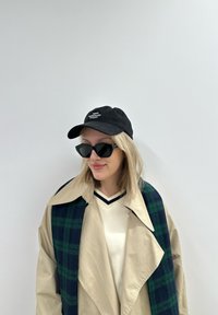Beige trench coat med stor krage, svart baseball caps med hvit logo, store svarte solbriller og grønn rutete skjerf drapert over skulderen.