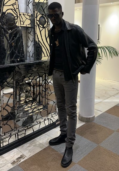 Chaqueta de cuero negra sobre una camisa negra, pantalones gris ajustados y botas negras. Presenta un objeto reflectante y una pared decorativa negra con una calavera.