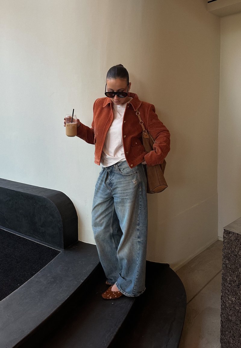 Femme portant une veste couleur rouille, une chemise blanche, un jean bleu ample et des chaussures marron, tenant un café glacé et un sac à main marron à l'intérieur près des escaliers.