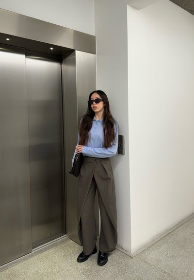 Mujer con gafas de sol y pantalones de pierna ancha de pie junto a la pared al lado de unas puertas de ascensor de acero inoxidable cerradas.