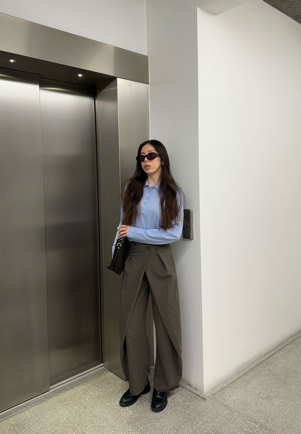 Femeie cu ochelari de soare și pantaloni evazați, stând lângă un perete, lângă ușile închise ale unui lift din oțel inoxidabil.