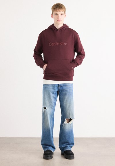 Sudadera color burdeos con bolsillo frontal y logo de Calvin Klein, combinada con jeans desgastados de color azul claro con grandes desgarros, usados con zapatos negros.