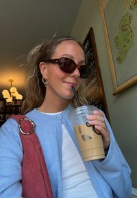 Cardigan azul-claro, blusa branca, óculos de sol tartaruga, brincos de argola dourados. Segurando um copo transparente com café gelado e um canudo preto.
