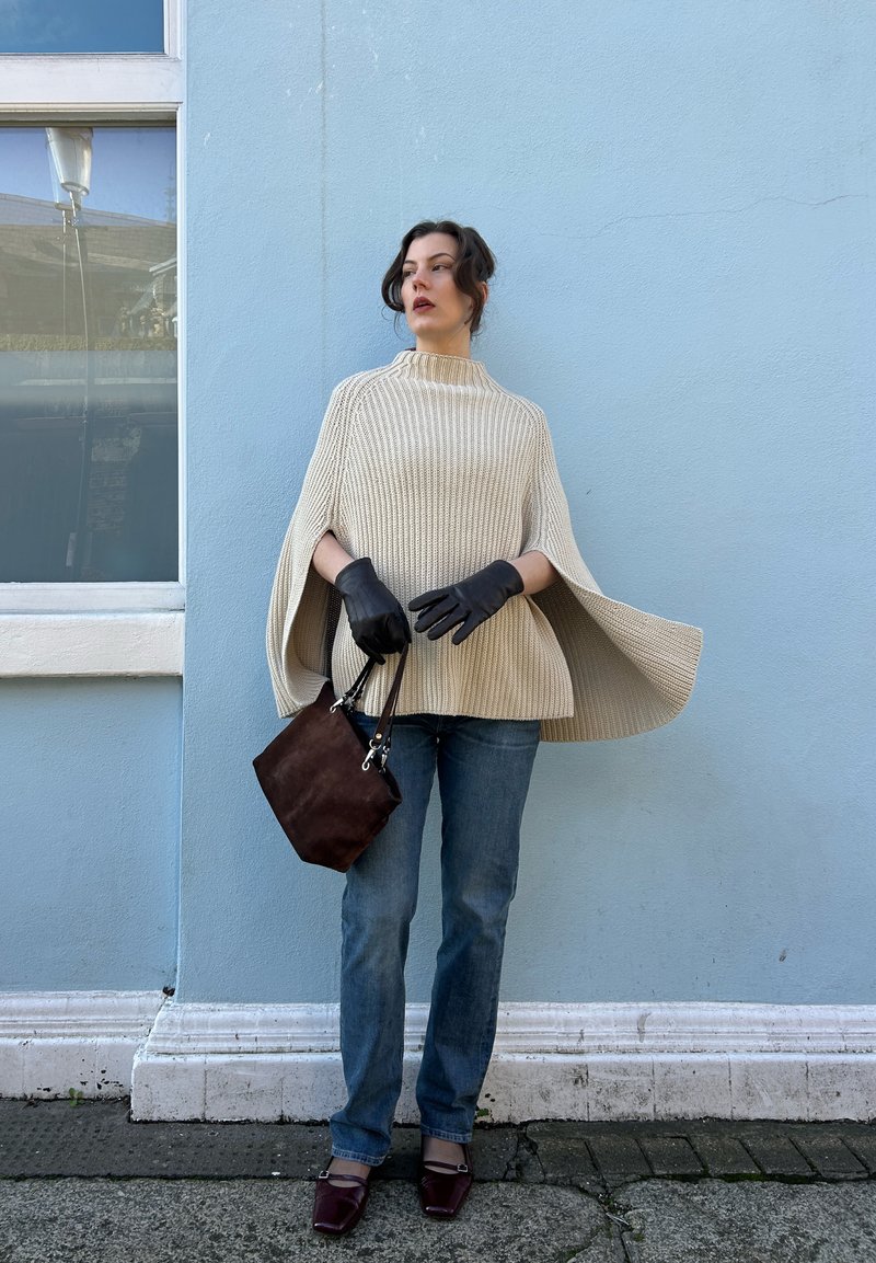 Femme debout contre un mur bleu, portant un pull cape beige, un jean bleu, des gants noirs, tenant un sac à main marron et chaussée de souliers bordeaux.