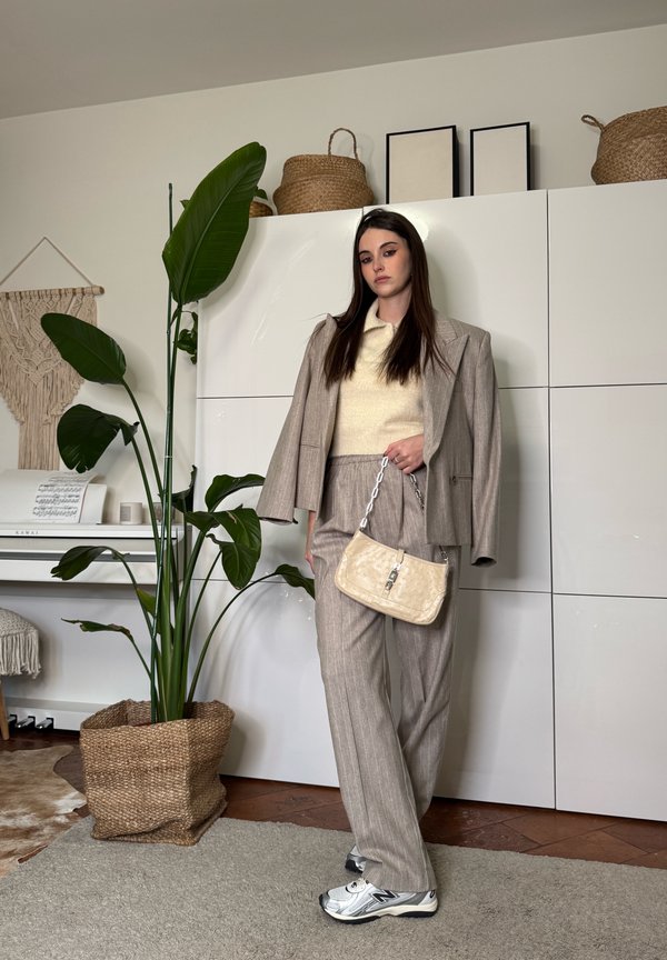 Donna con completo beige, maglione crema e sneakers, che tiene una piccola borsa, in piedi all'interno accanto a una pianta e a armadietti bianchi con ceste intrecciate.