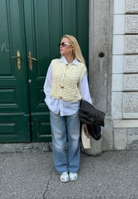 Dzianinowa kremowa kamizelka z dużymi guzikami noszona na białej koszuli, zestawiona z jasnoniebieskimi dżinsami i srebrnymi trampkami. Dodatki obejmują okulary przeciwsłoneczne i czarną kurtkę.