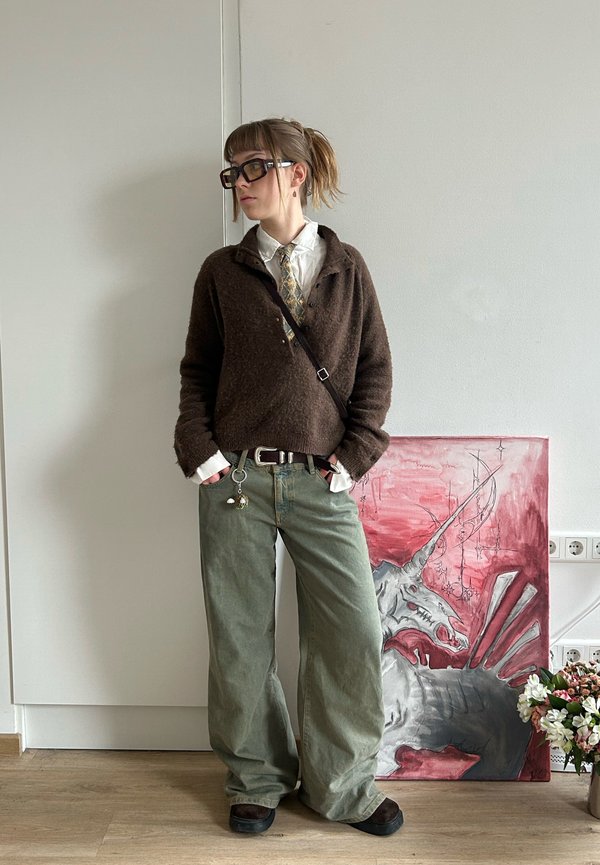 Osoba s okuliarmi, hnedý sveter, biela košeľa, vzorovaná kravata, zelené široké nohavice stojaca vedľa červeno-šedého obrazu jednorožca a kvetov.