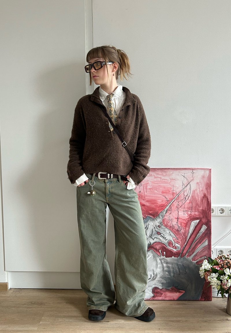 Persoană cu ochelari, purtând un pulover maro, cămașă albă, cravată cu model și pantaloni largi verzi, stând lângă o pictură de unicorn roșu și gri și flori.