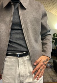 Szürke gyapjúdzseki gallérral, sötét pulóver fölött viselve. Fekete öv, világos színű nadrág és részletes tetoválások a kézen.