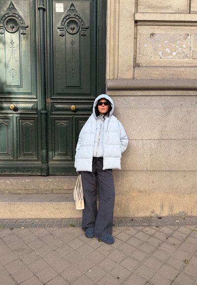 Persona con chaqueta acolchada blanca con capucha, gafas de sol oscuras, pantalones anchos a rayas, zapatos azules y sosteniendo una bolsa beige está de pie contra una pared de piedra.