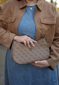 Brązowa torebka ze sztucznej skóry o zakrzywionym kształcie, z trójkątnym detalem logo i złotym okuciem. Modelka ma na sobie beżową kurtkę na lnianą sukienkę.