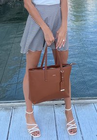 Καφέ δερμάτινη τσάντα tote με δύο χερούλια, χρυσά αξεσουάρ και ετικέτα. Ριγέ φούστα και λευκά σανδάλια με λουριά συμπληρώνουν το σύνολο.