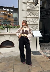 Kvinna som bär en beige kappa över en brun och gräddfärgad färgblockströja, svarta byxor med vida ben och vinröda skor, ståendes utanför en butik.