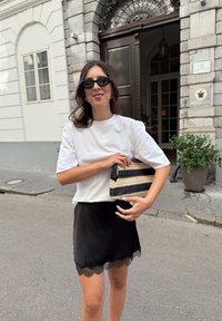 Bela majica s kratkimi rokavi, črna satenska krila z čipkastim robom, drži črno in kremasto črtasto pleteno torbico, stoji na mestni ulici.