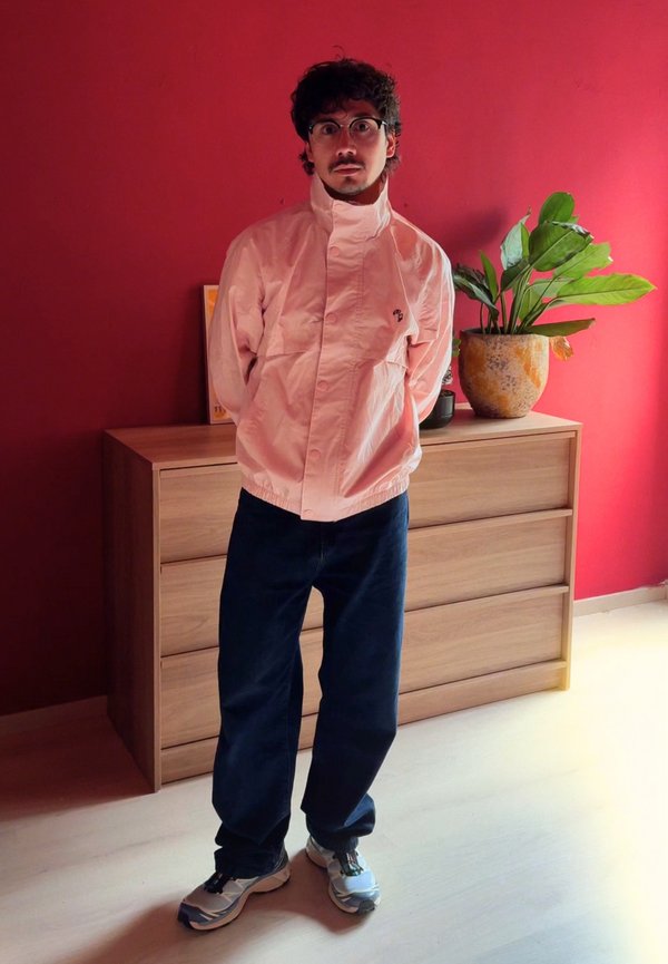Hombre con gafas que lleva una chaqueta rosa claro, pantalones oscuros y zapatillas, de pie frente a una cómoda de madera con una planta en maceta.