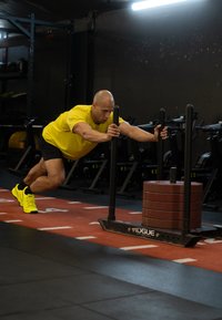 Rumena športna majica in črne kratke hlače, ki jih nosi oseba, ki potiska sani z rdečimi utežmi na telovadniškem tlaku, označenem z belimi črtami.