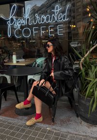 Μαύρο δερμάτινο μπουφάν με φούντες, κόκκινες κάλτσες, καφέ μοκασίνια, μαύρη τσάντα και ένα νέον σήμα "φρέσκος καβουρδισμένος καφές" στο φόντο.