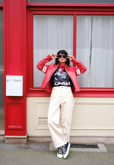 Chaqueta de cuero roja con rayas blancas, camiseta negra con gráfico, pantalones de pierna ancha beige, zapatillas negras y gafas de sol en forma de corazón contra un fondo rojo.
