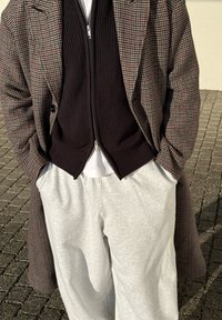 Personne portant un pardessus à carreaux, un pull zippé noir, une chemise blanche et un pantalon de survêtement gris clair, debout sur un sol pavé avec les mains dans les poches.