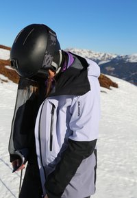 Veste de ski mêlant tissu blanc et noir, avec une fermeture éclair à l'avant, une capuche, et un design complexe mais discret. Portée avec un casque noir.