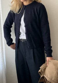 Cardigan tricoté bleu marine avec un col rond, fermeture à boutons et manches longues. Porté par-dessus une chemise blanche et un pantalon noir.