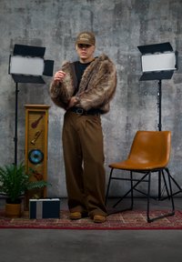 Jeune personne portant une veste en fourrure marron et une casquette, posant entre deux éclairages de studio près d'une chaise, d'une plante, d'une radio vintage et d'une vitrine musicale.