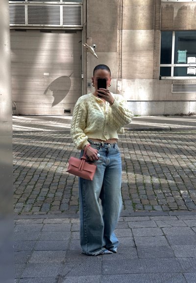 Cárdigan de punto trenzado crema con botones, vaqueros de mezclilla azul anchos y un pequeño bolso rosa. Texturas suaves y ajuste relajado.