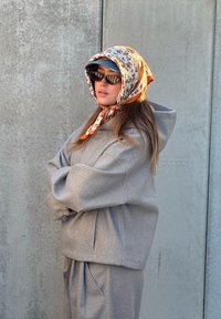 Femme portant un foulard orange à motifs floraux sur une casquette grise, des lunettes de soleil noires, un sweat à capuche gris et un pantalon assorti, debout contre un mur en béton.